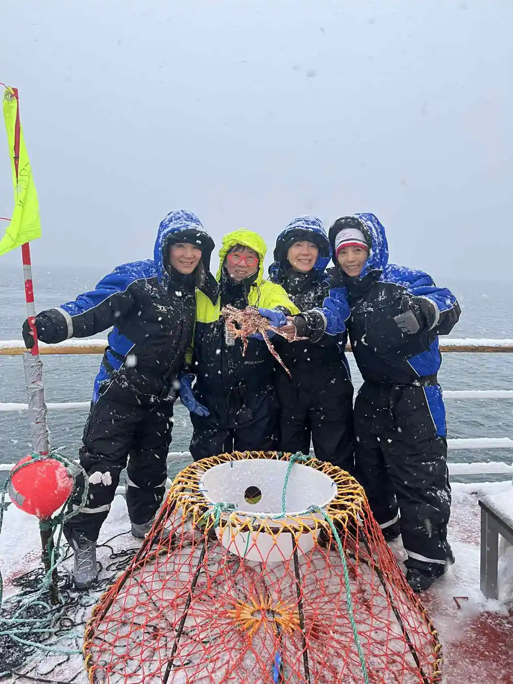 Five guests in insulated suits crowded around a crab pot on deck in snow