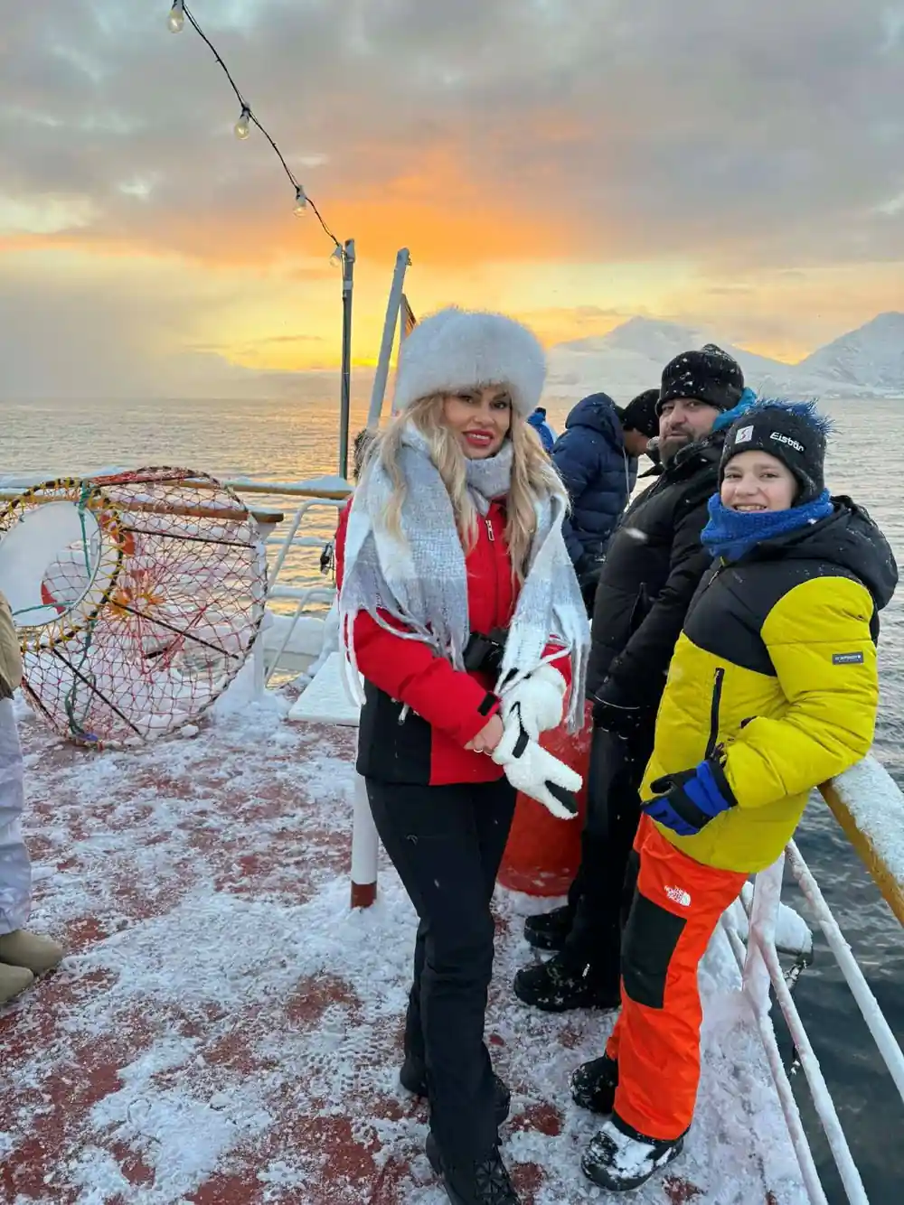 Guests beside a crab pot on deck at sunset with snowy peaks behind